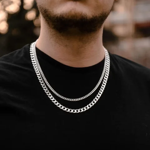 Dvoslojna kombinacija muških srebrnih ogrlica na crnoj majici — tanji Cuban Link lanac uz vrat i deblji, duži sloj ispod, u razmaku od 5 cm.