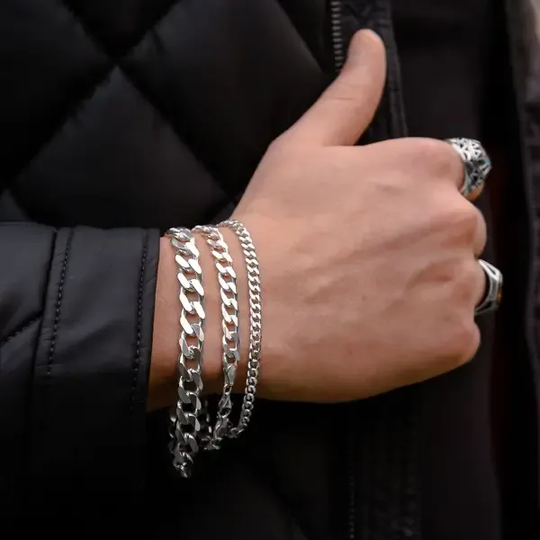 Tri srebrne Cuban Link narukvice, S, M i L, zajedno na muškom zglobu uz crnu jaknu.