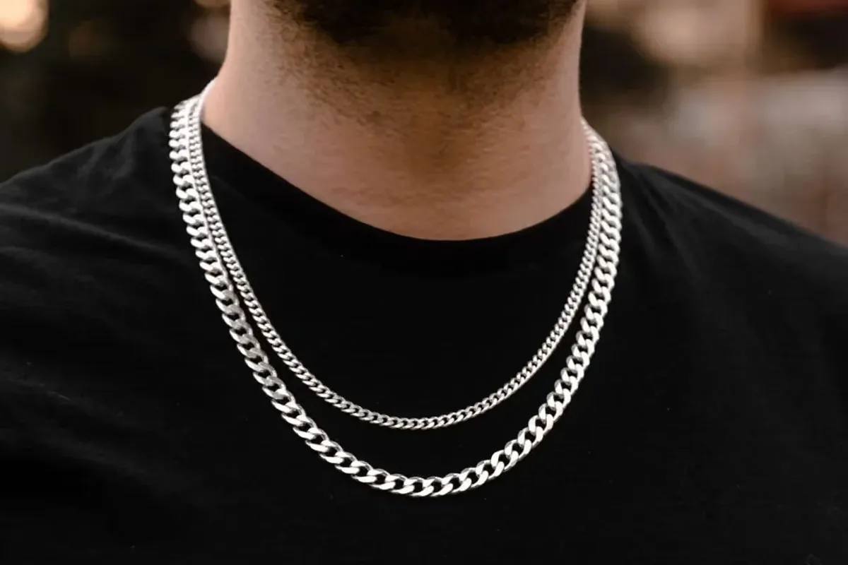 Dve muške srebrne ogrlice pancir rada (Cuban Link) različite debljine, slojevito nošene na crnoj majici.