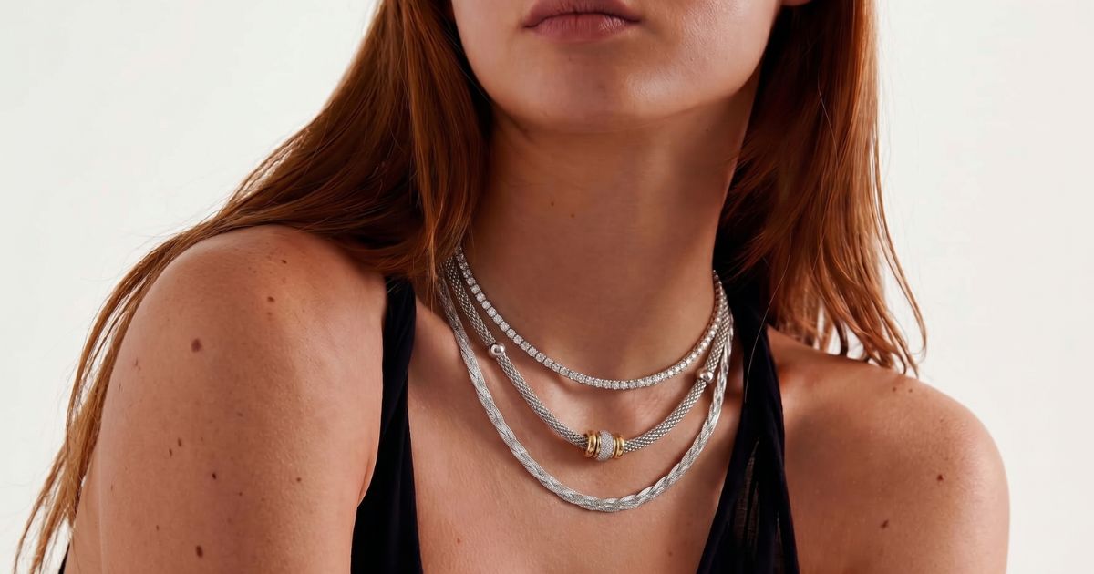 Žena nosi tri srebrne ogrlice različitih dužina preko bele košulje, prikazujući layering trend.